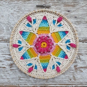 Könnte beinhalten: Ein farbenfrohes gehäkeltes Mandala mit einem Sternburst-Design in den Farben Pink, Gelb, Blau und Grün. Das Mandala ist in einem Holzreifen gerahmt und mit kleinen weißen Perlen verziert.