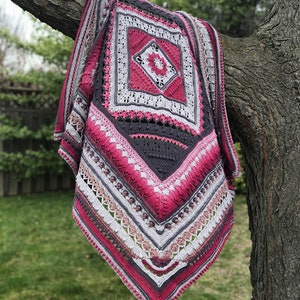 May include: A crocheted blanket with a pink, gray, and white color scheme. The blanket is hanging on a tree branch and has a square design in the center.