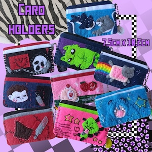 May include: Nine felt card holders with various embroidered designs. The designs include a green cat, a rainbow with a pink toast character, a red heart with a white skull, a red and black design with a white skull, a blue design with a white character, a pink design with a white character, a red design with a white character, a pink design with a green character, and a blue design with a white character. The card holders are 7.5 cm x 10.2 cm.