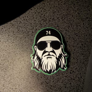 May include: A sticker featuring a stylized illustration of a bearded man wearing sunglasses and a cap with the number 74. The design has a black and white color scheme with green accents.