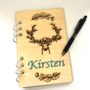 May include: A wooden spiral bound journal with a woodburned design of a deer with floral antlers. The name "Kirsten" is written in turquoise lettering on the front cover.