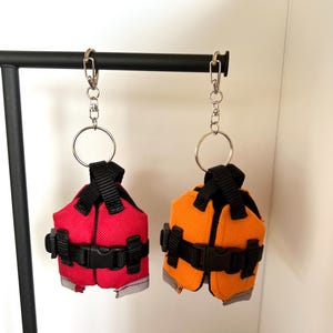 May include: Two miniature life jackets with keychains. One is red and the other is orange. Both have black straps and buckles.