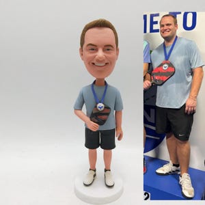 May include: A bobblehead figurine of a man in a light blue top, black shorts, and white trainers. The figurine has a blue lanyard with a paddle-shaped pendant. The image also shows a real person wearing a similar outfit and holding a paddle.