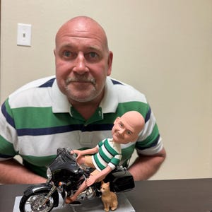 May include: A figurine of a person riding a black motorbike with a cat sitting on the side. The figurine is on a grey base.