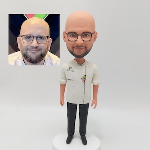 May include: A custom-made caricature figurine of a person wearing a white chef's uniform with black trousers and black shoes. The figurine has a large head and a small body. The person is wearing glasses and has a beard. The figurine is standing on a white base.