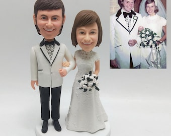 May include: A wedding cake topper featuring a smiling bride and groom figurine. The groom wears a white tuxedo with black pants and a bow tie. The bride is in a white lace dress and holds a bouquet. A framed wedding photo is in the background.