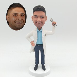 May include: A custom bobblehead of a smiling man in a white lab coat, blue shirt, and dark blue pants, holding a dental tool. The bobblehead is on a white base. An oval portrait of the man is in the upper left corner.