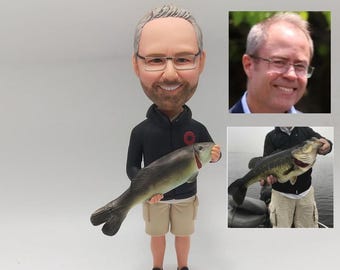 May include: A custom bobblehead figurine of a person holding a fish. The figurine is wearing a black jacket, khaki shorts, and black shoes. The person has gray hair, a beard, and is wearing glasses.