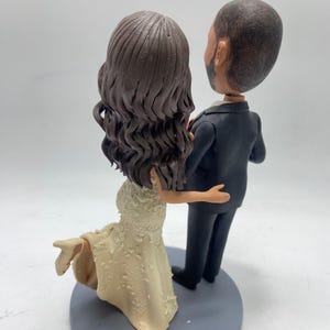May include: A wedding cake topper featuring a bride and groom figurine. The bride is wearing a white dress and has long brown hair. The groom is wearing a black suit and has short brown hair.
