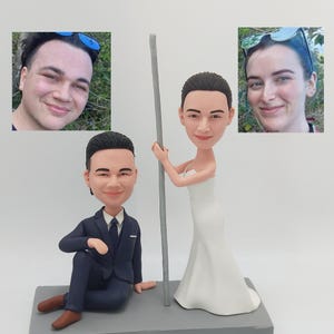 May include: A custom-made figurine wedding cake topper featuring a bride and groom. The bride is wearing a white dress and holding a silver pole. The groom is wearing a dark blue suit and sitting down. The figurines are made to look like real people.