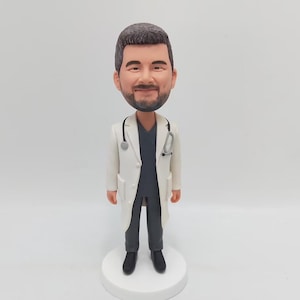 May include: A custom bobblehead figurine of a doctor wearing a white lab coat with a stethoscope around their neck. The doctor has a black undershirt and black pants. The bobblehead is standing on a white base.