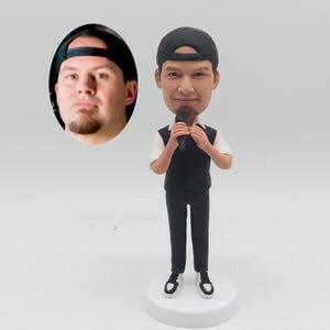 May include: A custom bobblehead figurine of a man wearing a black cap, white shirt, black waistcoat, and black trousers, holding a microphone. The figurine stands on a white base. An oval portrait of a man wearing a black cap is in the background.