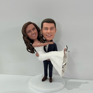 May include: A wedding cake topper featuring a bride and groom figurine. The groom is holding the bride in his arms. The bride is wearing a white dress and the groom is wearing a dark suit.