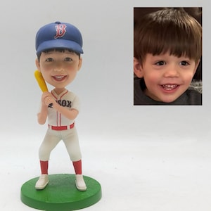 Puede incluir: Una figura de bobblehead personalizada de una persona sonriente con uniforme de béisbol, sosteniendo un bate amarillo. La figura lleva una gorra de béisbol azul con el logo "B" rojo, una camiseta blanca con "SOX" en el pecho y calcetines rojos. La figura está sobre una base verde.
