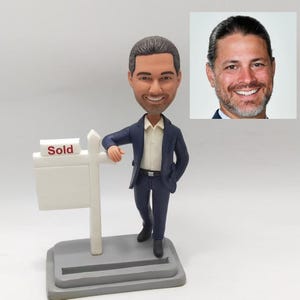 May include: A custom bobblehead figurine of a person in a blue suit, standing next to a white real estate sign with the word "Sold" on it. The figurine has a realistic face and is holding a hand to their chin.