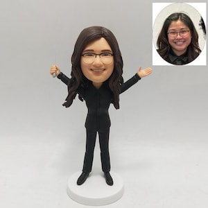 May include: A custom bobblehead figurine of a person with long brown hair, glasses, and a black suit, holding a small silver object. The figurine is on a white base, with a smaller photo of the person in the upper right corner.