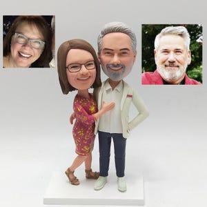 May include: A custom-made figurine set of a couple. The woman is wearing a pink and gold dress and the man is wearing a white shirt and a white blazer. They are both smiling and standing close together.