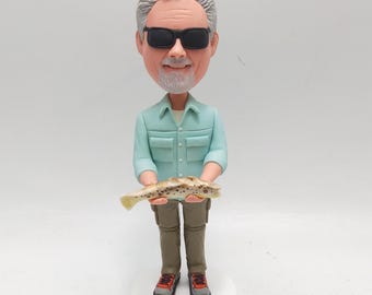 May include: A figurine of a man wearing sunglasses, a light blue shirt, and khaki pants, holding a fish. The figurine has a bobblehead design and is standing on a white circular base. The man's shoes are black, red, and gray.