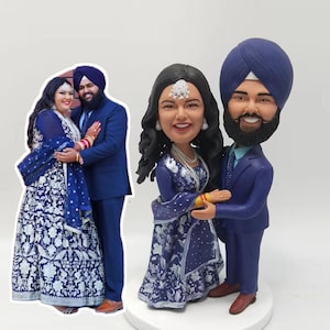 Op de afbeelding: Een op maat gemaakt beeldje van een stel in traditionele Indiase trouwkledij. De bruid draagt een blauwe en witte jurk met bloemenborduurwerk, en de bruidegom draagt een blauw pak met een tulband. De beeldjes zijn in een romantische omhelzing geposeerd.