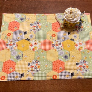 May include: A yellow and green quilted placemat with a honeycomb pattern and bees. The placemat has a variety of floral and geometric patterns. The text "Bee Happy" is on a mug in the image.