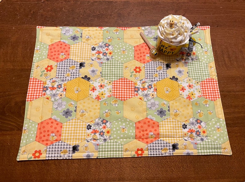 May include: A yellow and green quilted placemat with a honeycomb pattern and bees. The placemat has a variety of floral and geometric patterns. The text "Bee Happy" is on a mug in the image.