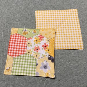 May include: Two quilted fabric coasters. One coaster is yellow with a white gingham pattern. The other coaster is a patchwork of yellow, green, red, and white gingham fabric with a floral and bee pattern.