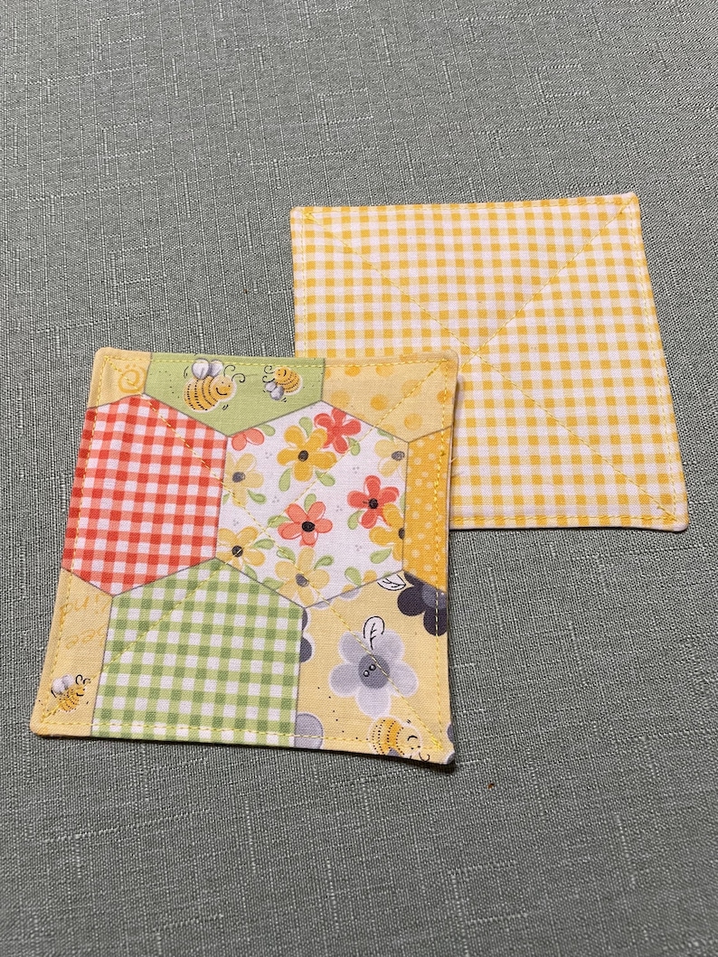 May include: Two quilted fabric coasters. One coaster is yellow with a white gingham pattern. The other coaster is a patchwork of yellow, green, red, and white gingham fabric with a floral and bee pattern.