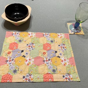 May include: A colorful patchwork placemat with a hexagonal pattern in shades of yellow, green, pink, and white. The placemat is made of fabric and has a quilted texture.