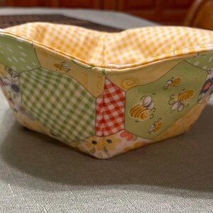 May include: A yellow, green, and red fabric bowl with a honeycomb pattern and bees. The bowl is made of patchwork fabric and is perfect for holding small items.