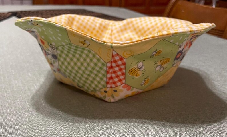 May include: A yellow, green, and red fabric bowl with a honeycomb pattern and bees. The bowl is made of patchwork fabric and is perfect for holding small items.
