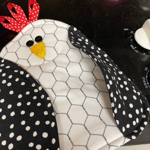 May include: A white and black polka dot chicken-shaped pot holder with a red and white polka dot bow on top. The pot holder has a chicken wire pattern on the front.