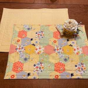 May include: Two yellow and white placemats with a honeycomb pattern. The top placemat is a simple gingham pattern. The bottom placemat has a patchwork design with bees, flowers, and polka dots. A mug with the text "bee happy" is sitting on the placemats.
