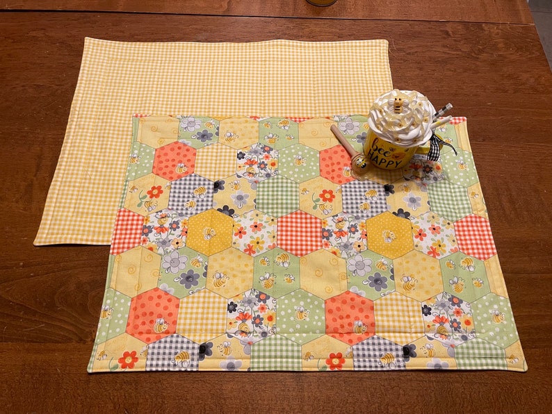 May include: Two yellow and white placemats with a honeycomb pattern. The top placemat is a simple gingham pattern. The bottom placemat has a patchwork design with bees, flowers, and polka dots. A mug with the text "bee happy" is sitting on the placemats.