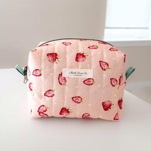 May include: A pink quilted cosmetic bag with a strawberry pattern. The bag has a black zipper, mint green handles, and a white label that reads "Mochi Luv Co."