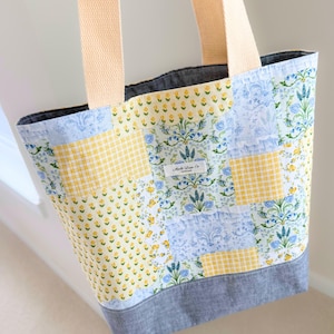 Peut inclure: Un sac fourre-tout patchwork avec un motif floral et vichy jaune et bleu. Le sac a des sangles beiges et un fond gris. L'étiquette indique "Made Lane Co."