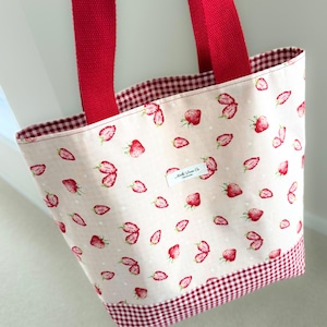 Peut inclure: Un sac fourre-tout rouge et blanc avec un imprimé de fraises et une bordure en vichy rouge. Le sac a des anses en tissu rouge et une étiquette blanche avec le texte "Make Space Co."