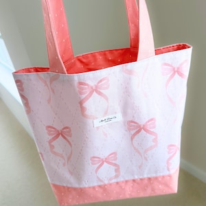 May include: A pink tote bag with a repeating pattern of pink bows on a white background. The bag has pink handles and a pink polka dot trim. The interior is a solid coral color. A small label with text is visible on the front.