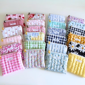 May include: A collection of quilted cosmetic bags in various patterns and colors, including floral, gingham, and daisy prints. Each bag features a zipper closure and a small label. The bags are arranged on a white surface.