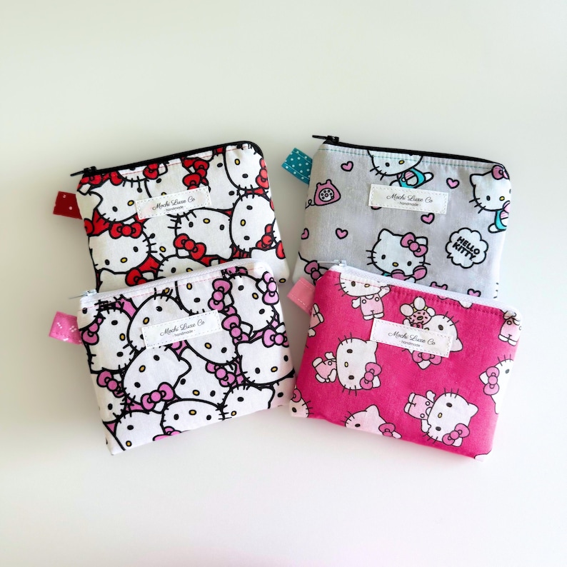 May include: Four small zippered pouches with Hello Kitty designs. The pouches are made of white fabric with pink, red, and blue accents. The pouches are all different designs, but they all feature Hello Kitty and the text "Hello Kitty".