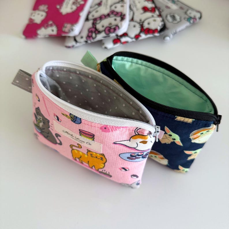 May include: Two zippered pouches, one pink with a cat and sushi design and the text "Mocha Llama Co." and the other navy blue with a green alien design.