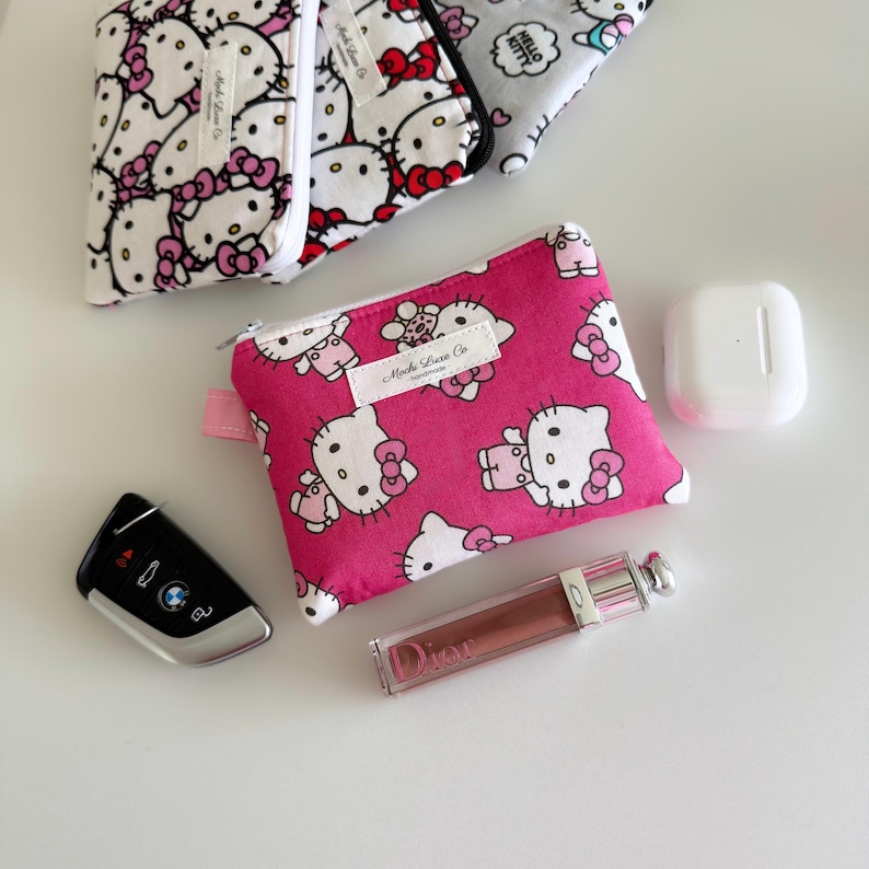 May include: A pink zippered pouch with a white background and a repeating pattern of Hello Kitty characters. The pouch is made by "Mocha Love Co."