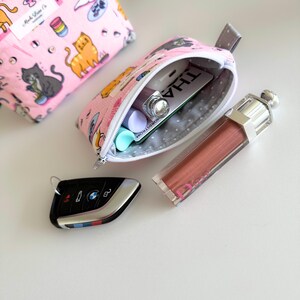 May include: A pink zippered pouch with a cat print design. The pouch is open and contains a lip gloss, a credit card, and a small tube of lip balm. A car key is visible in the foreground.