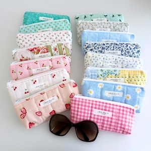 May include: A collection of rectangular quilted fabric pencil cases in various floral, fruit, and gingham patterns, each featuring a white label with the text Mochi Luxe Co. A pair of dark brown sunglasses rests in front of the pouches.