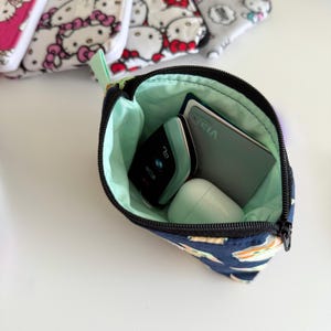 May include: A blue and white patterned zippered pouch with a green lining. The pouch is open and contains a black electronic device, a white earbud case, and a grey credit card with the word "VISA" printed on it.
