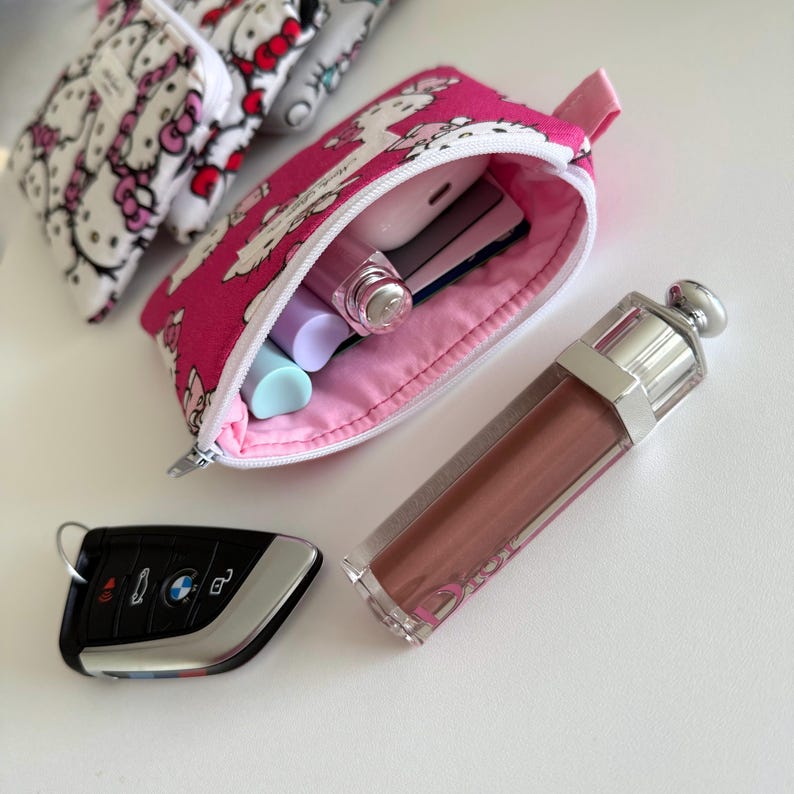 May include: A pink zippered pouch with a Hello Kitty print design. The pouch is open and contains various items, including a pink lip gloss, a white and silver phone charger, and a blue and white lip balm.