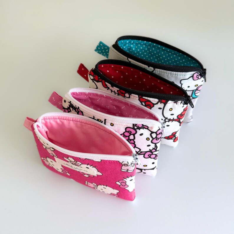 May include: Four Hello Kitty themed zippered pouches in different colours. The pouches are lined with a white fabric with small white polka dots. The pouches are open and the zippers are visible.