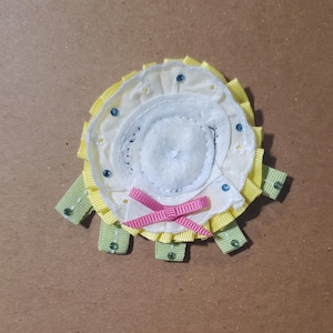Peut inclure: Une broche fleur en tissu faite à la main avec un centre blanc, entourée de couches jaunes et crème. La broche comporte un nœud en ruban rose et de petits ornements bleus. Des accents de ruban vert s'étendent de la base.