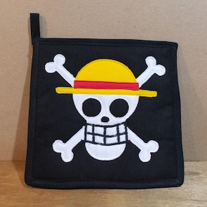 May include: Black pot holder with a white embroidered skull wearing a yellow and red hat with crossed bones.
