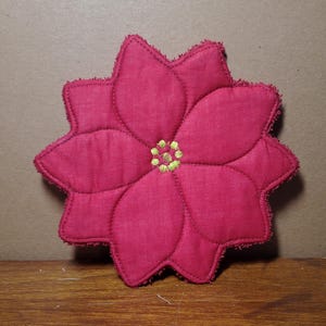 Puede incluir: Un posavasos acolchado en forma de flor de pascua magenta, con un centro bordado en amarillo. El posavasos tiene un borde festoneado y detalles cosidos, perfecto para la decoración navideña. El posavasos es de tela.
