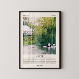 May include: Framed art print featuring a scenic view of Odense, Denmark. The image shows a river with people on paddleboards and a cyclist on a bridge. The print has text at the top and bottom, with the word "Odense" prominently displayed.